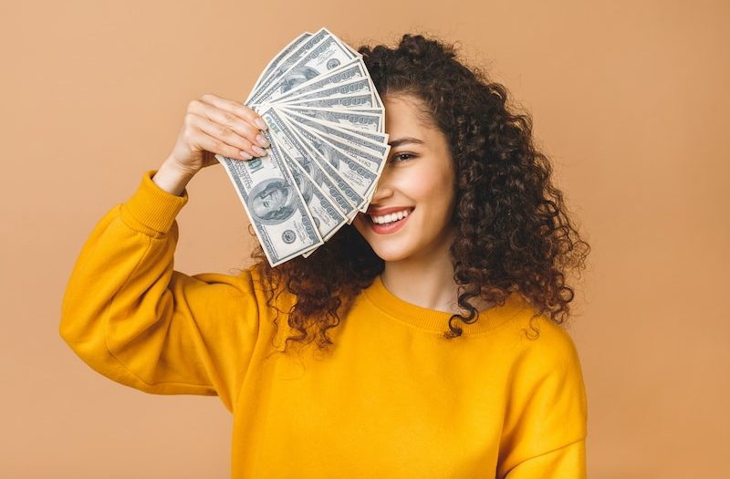 Portrait of a cheerful young woman holding money banknotes and celebrating isolated over beige background.