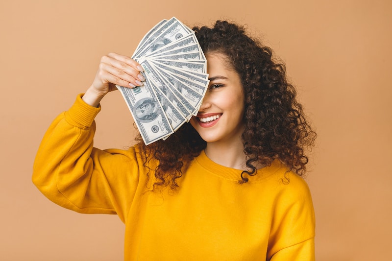 Portrait of a cheerful young woman holding money banknotes and celebrating isolated over beige background.